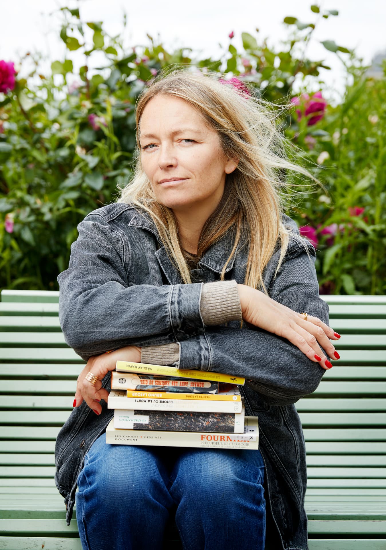 Maud Zilnyk assise sur un banc, bras croisés sur une pile de livres, devant un rosier en fleurs